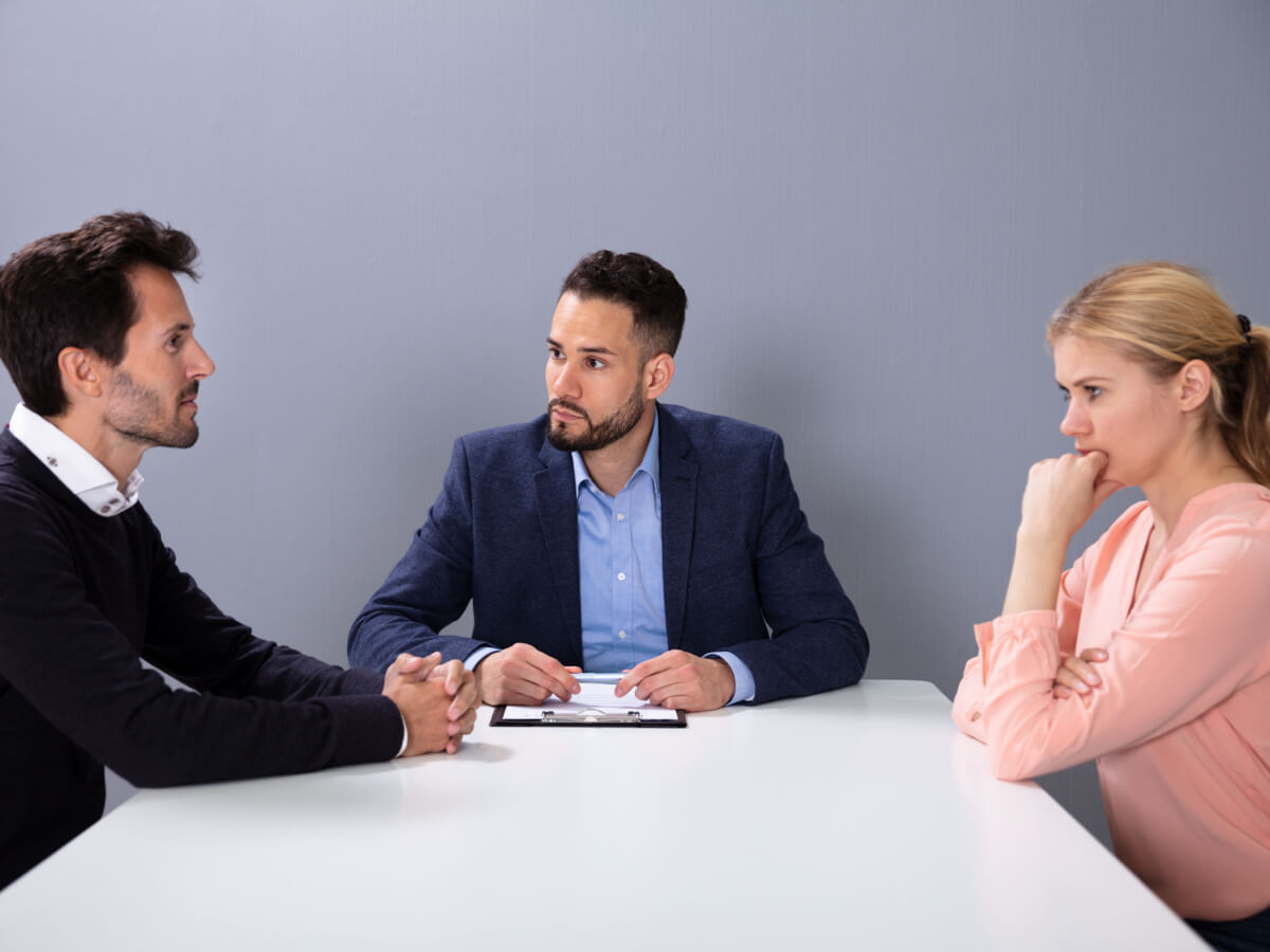 A couple sitting a table across from one another with a lawyer between them, tense expressions on their faces