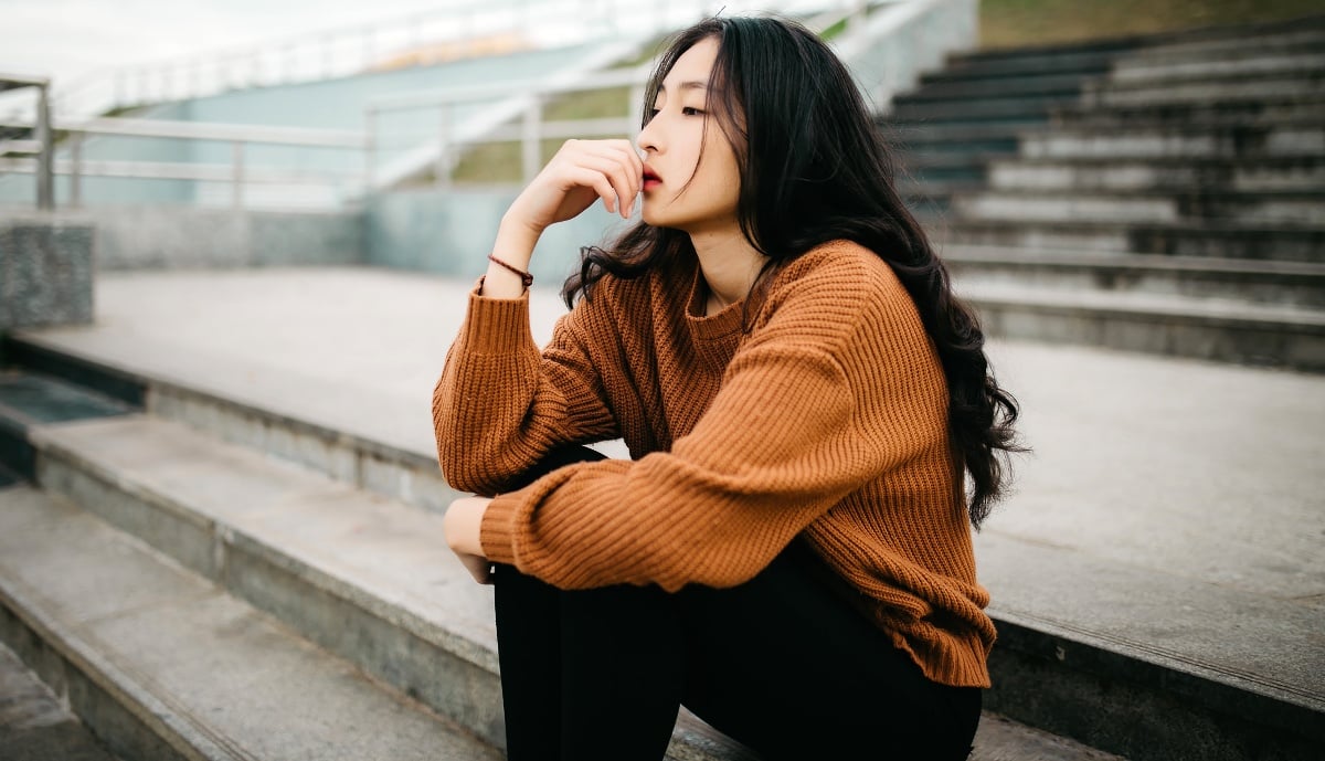 Girl wearing an orange sweater, sitting on bleachers with a contemplative expression.